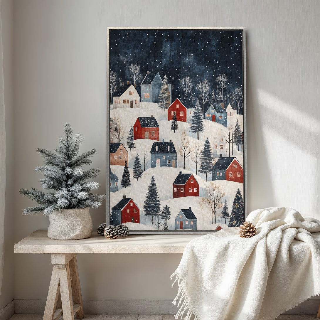 Framed Wall Art of a winter village scene in a folk art style. Features a snowy landscape with red and blue houses, set against a dark blue sky.