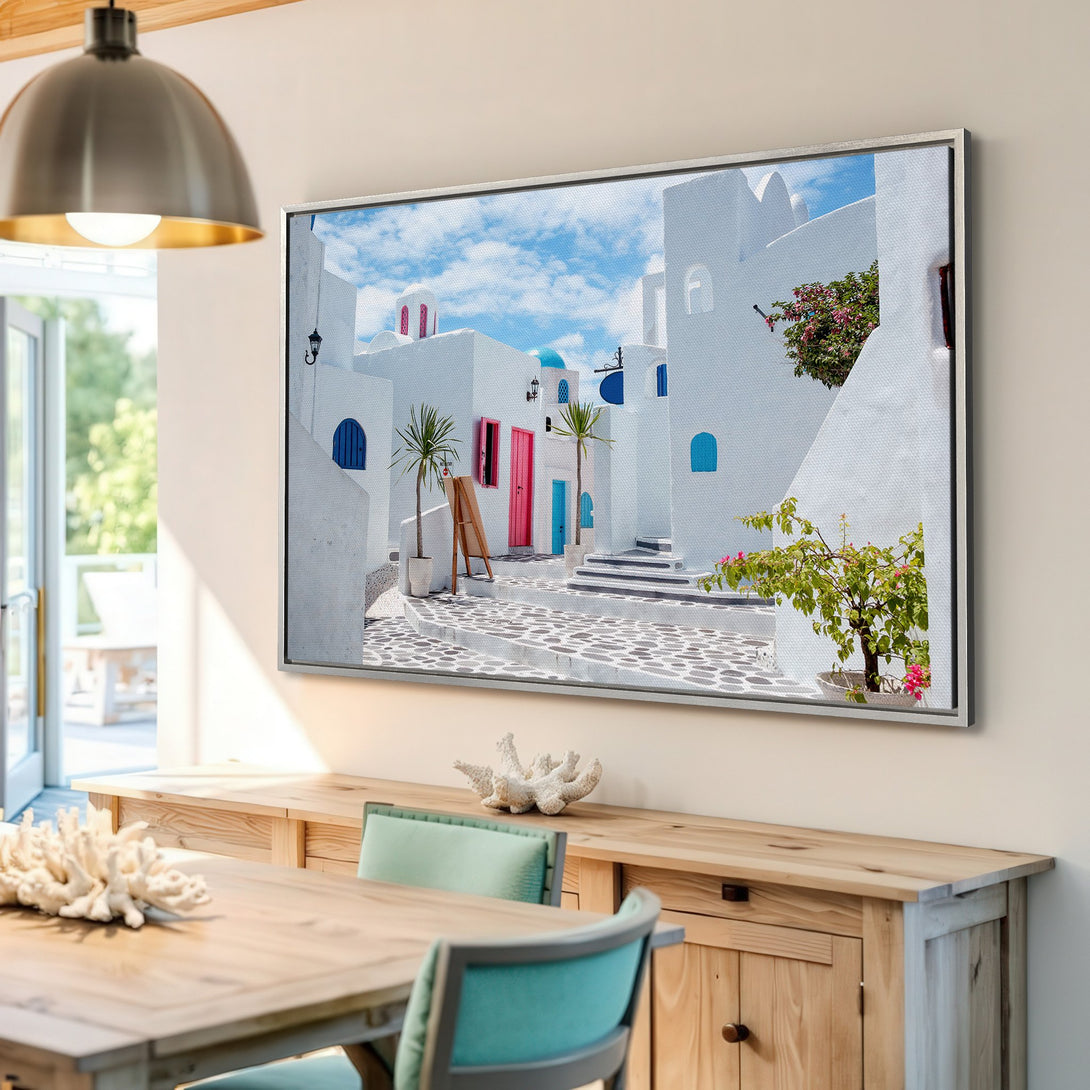 Framed Wall Art: Santorini street scene canvas print. White buildings, blue accents, and pink flowers. Bring Mediterranean vibes to your home!