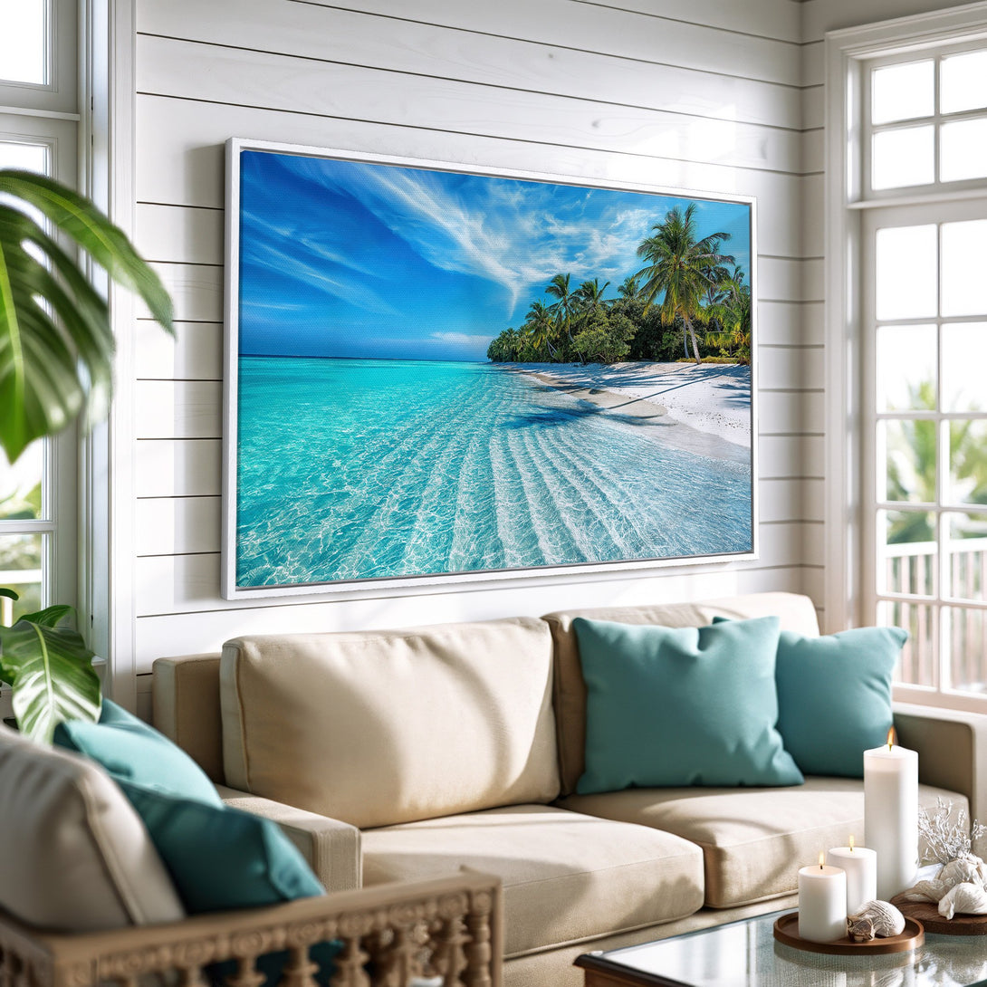 Framed Wall Art Canvas Print of tropical beach scene with blue water and white sand. Coastal decor in a bright, airy living room with teal and beige accents.