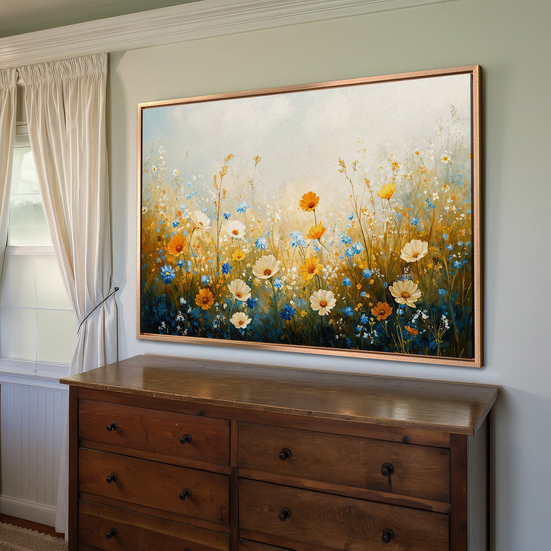 Framed Wall Art featuring a vibrant floral field painting. Warm yellow, blue, and white flowers pop against a soft, light blue and gold background. Adds a touch of nature.